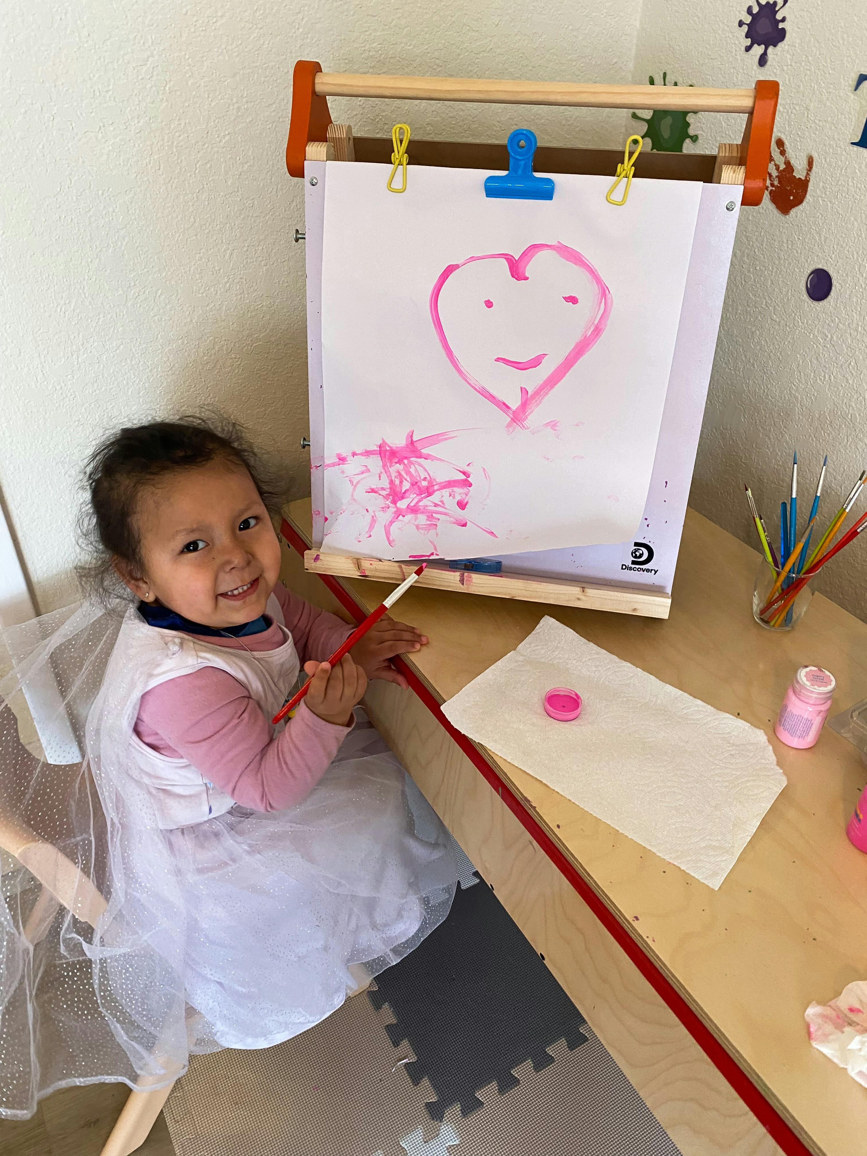 Child in a white dress painting a pink heart on an easel