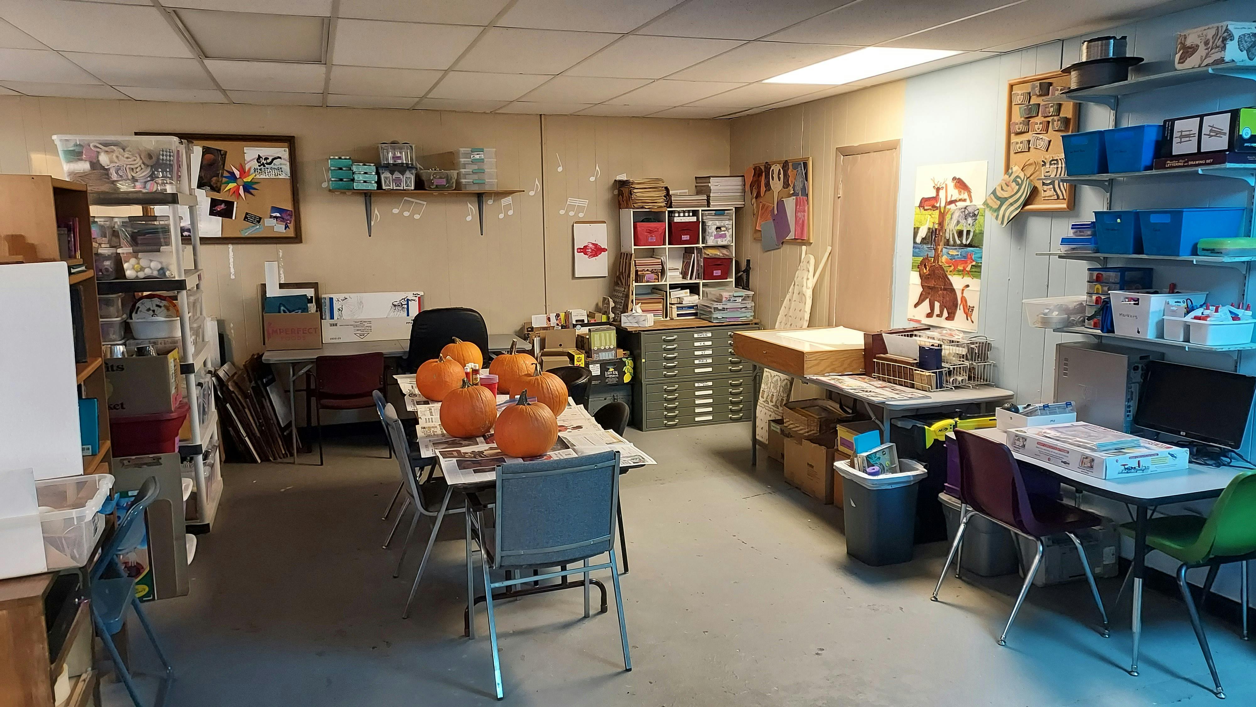A cluttered art room with tables, chairs, shelving, and pumpkins on a table