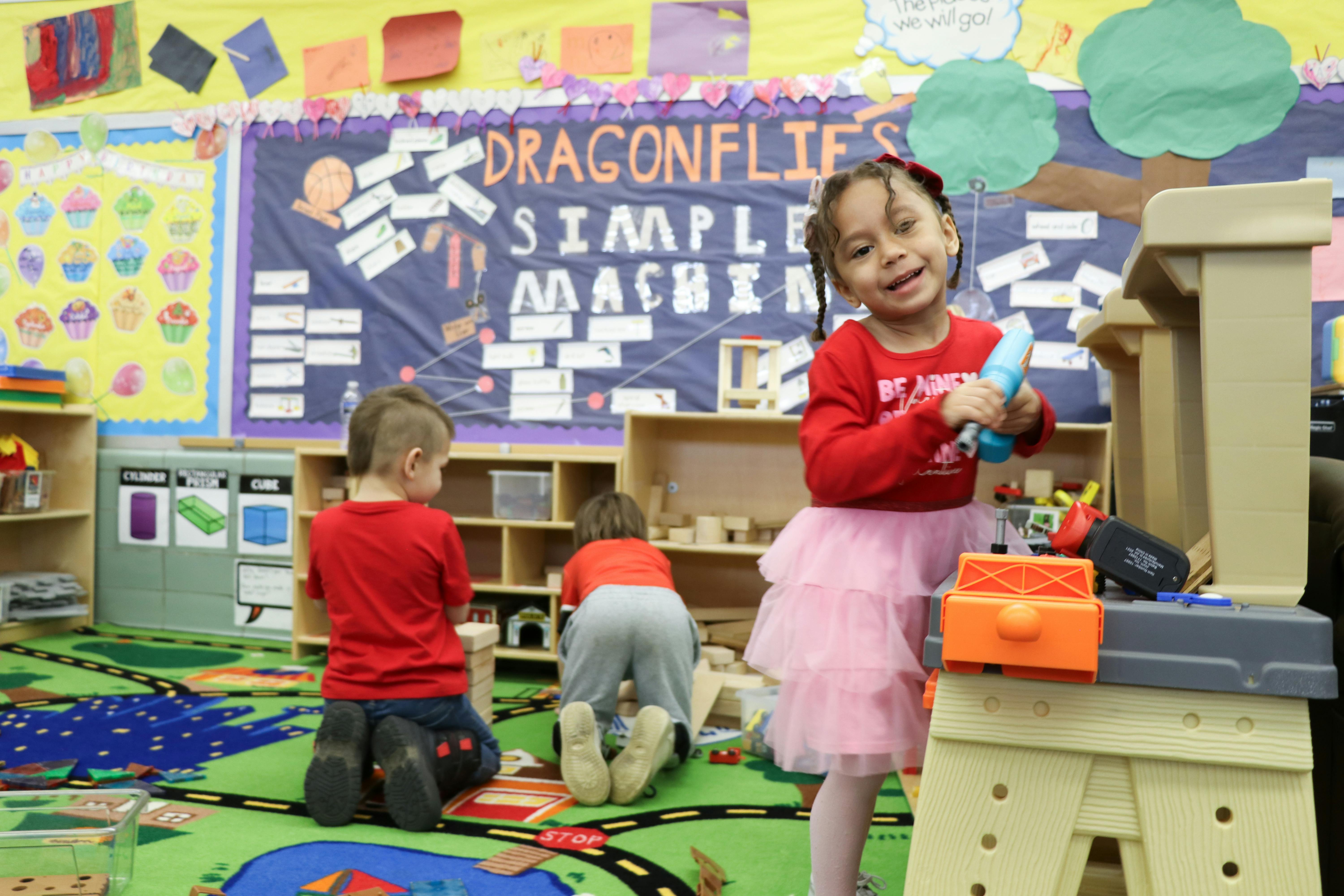 Child in pink skirt holding toy, two children playing in background