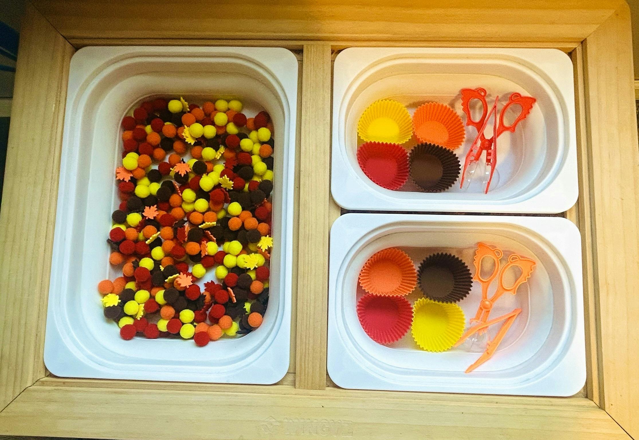 Craft supplies in white bins, including pom-poms, cups, and scissors