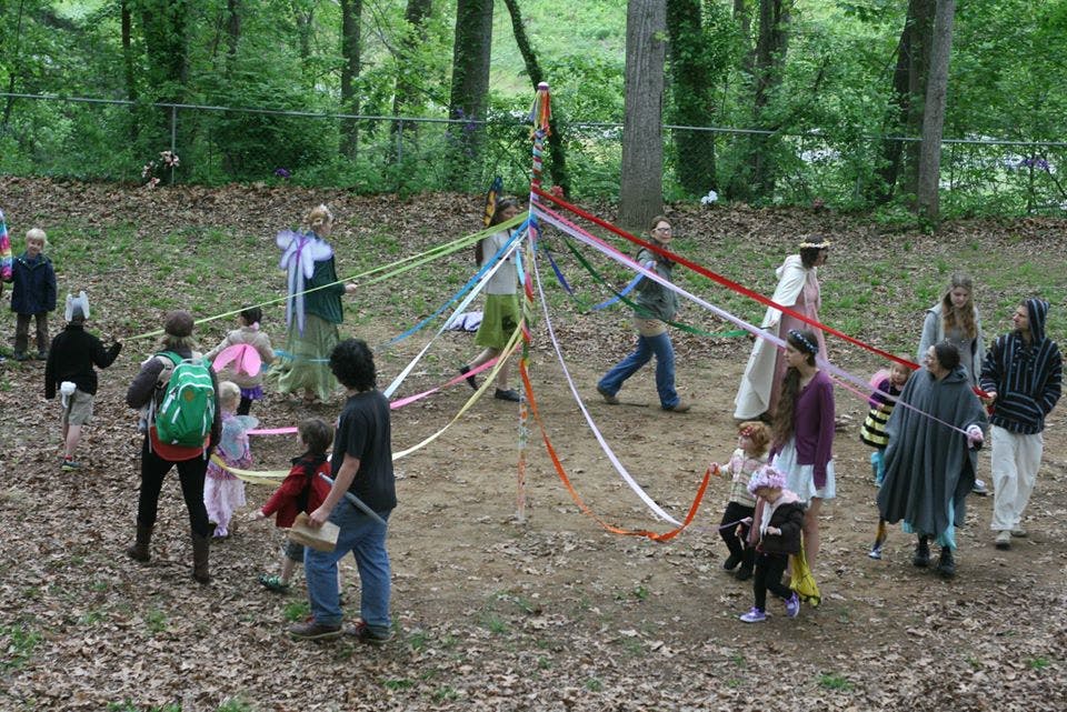 People and children dancing around a maypole with colorful ribbons in a wooded area