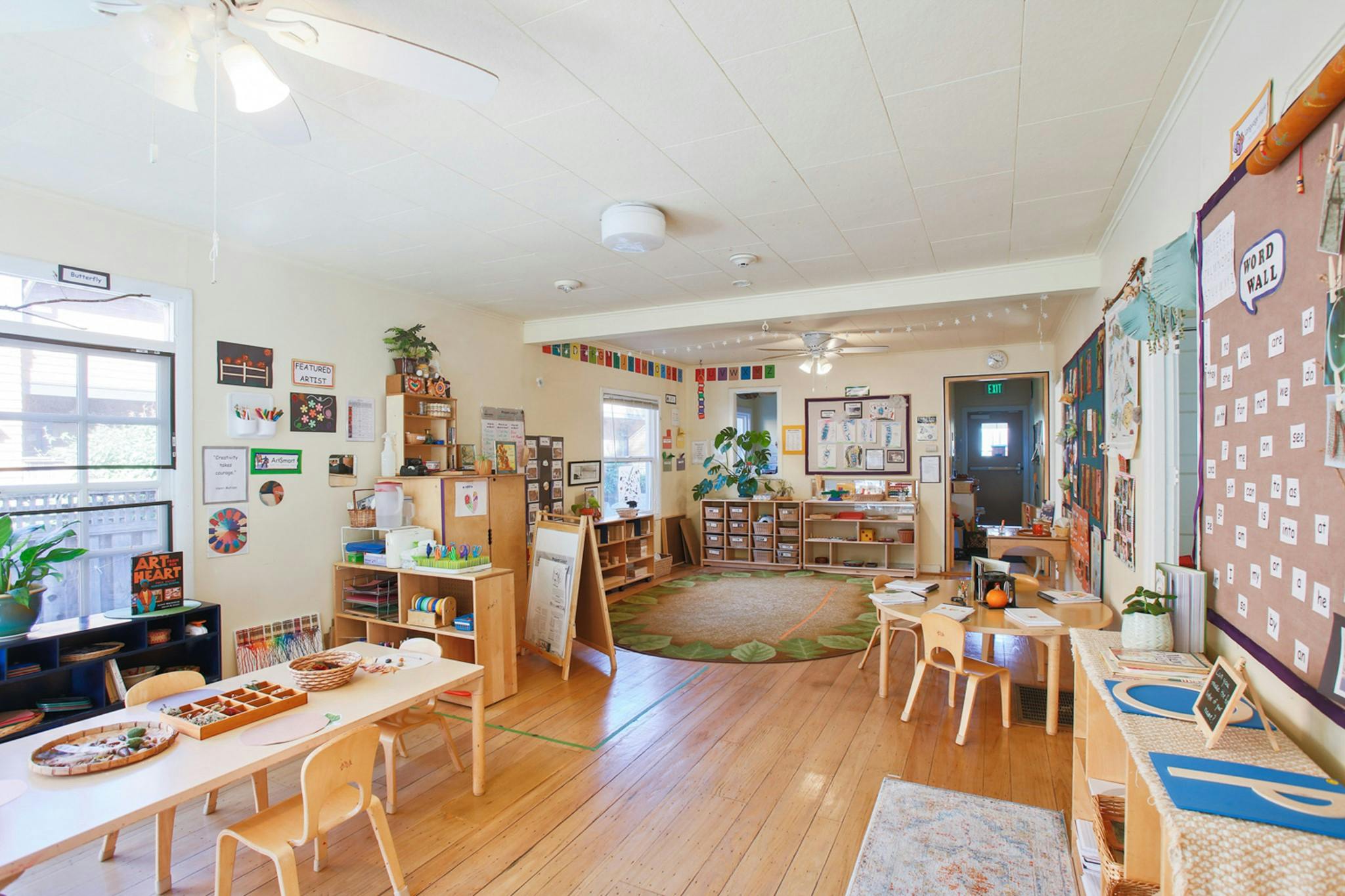 A classroom with wooden tables and chairs, shelves, and a circular rug