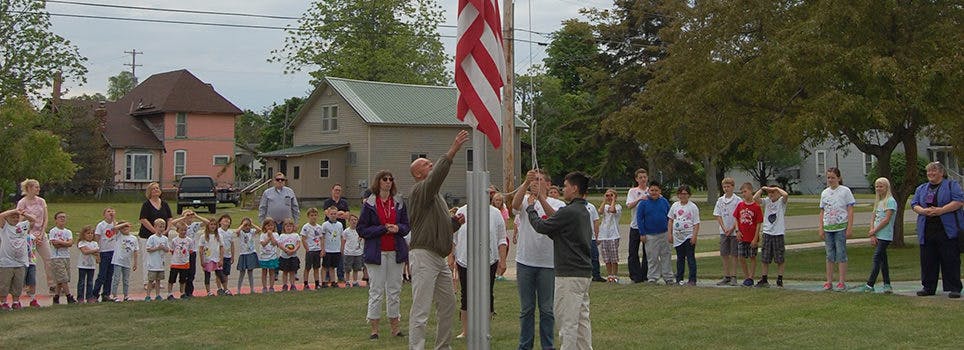People raising an American flag on a flagpole with children watching
