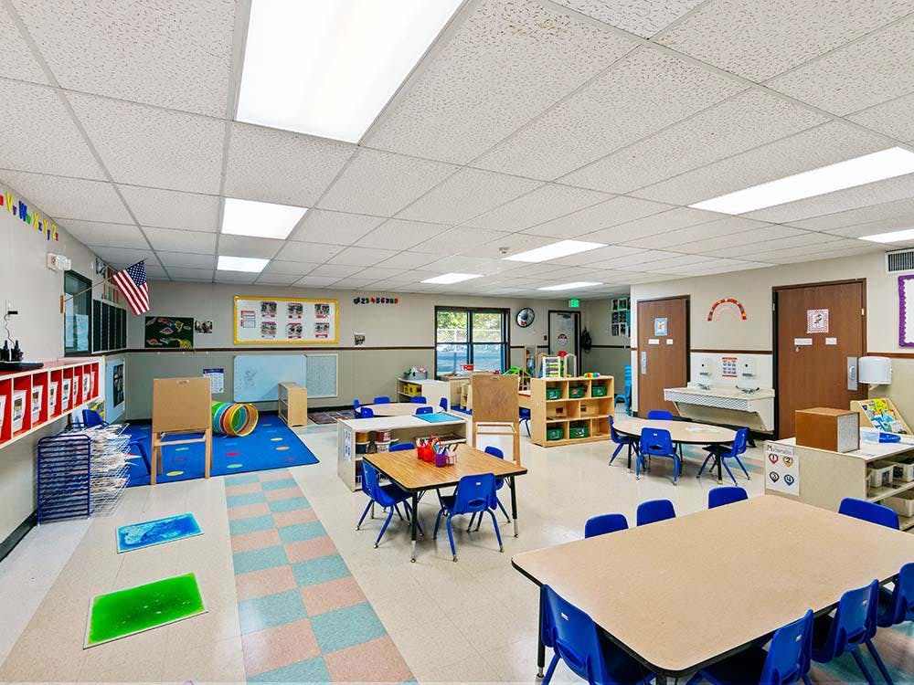 Empty classroom with tables and blue chairs set up, activity rug, and supplies