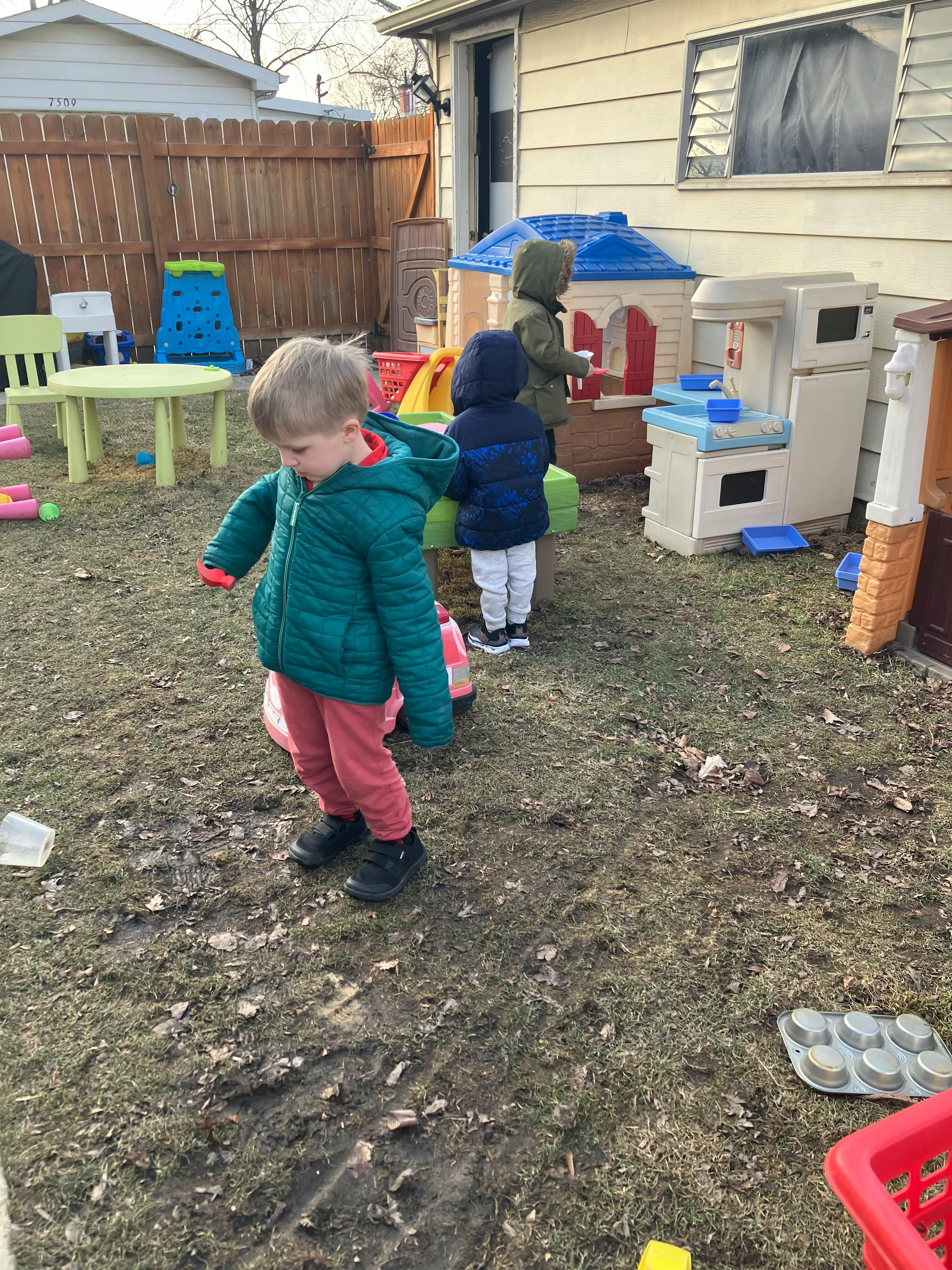 Three children playing outdoors next to toy playhouses and a play kitchen