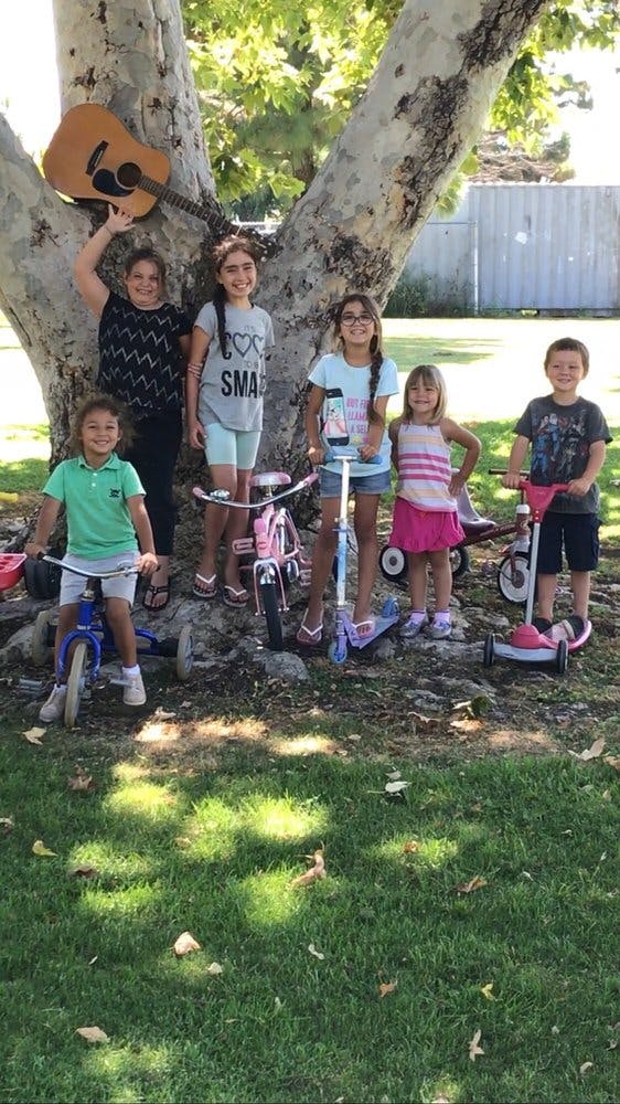 Group of children with bikes, scooters, and a guitar under a tree