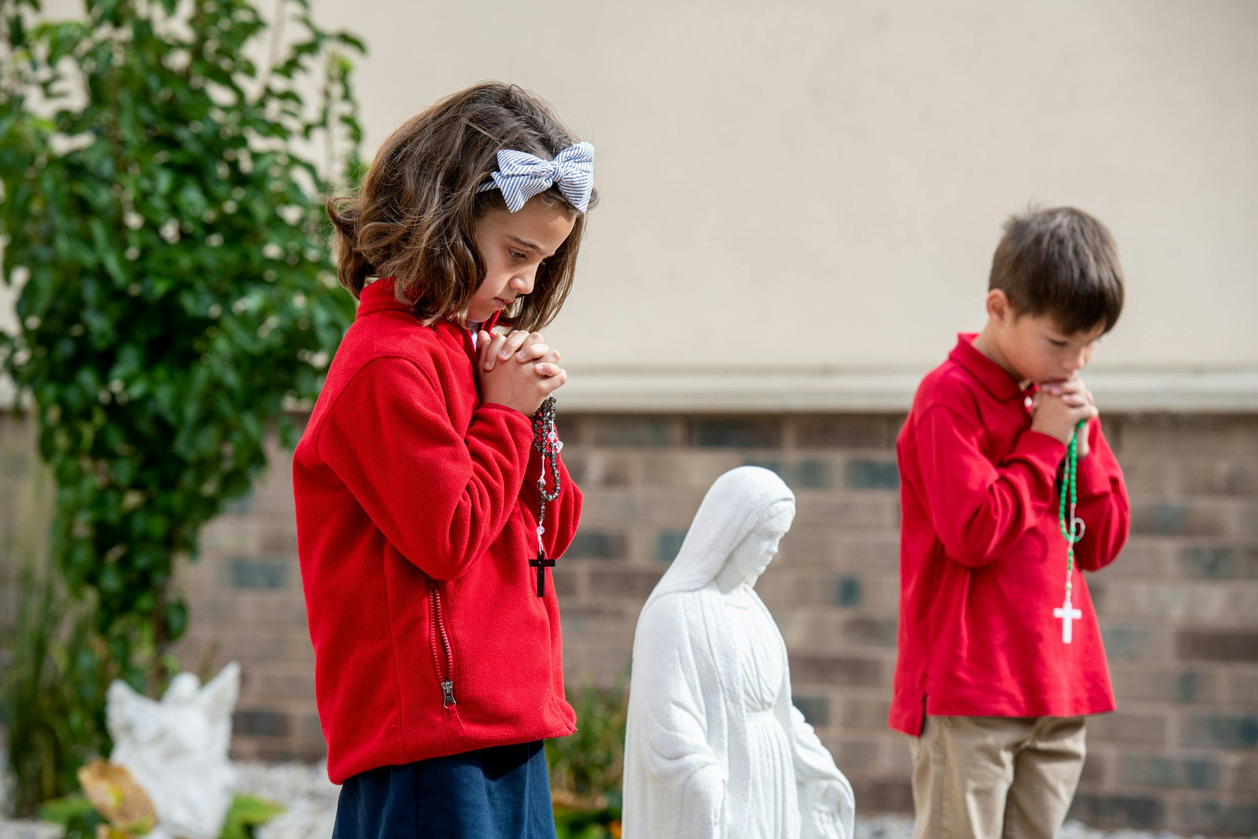 Two children praying with rosaries next to a statue