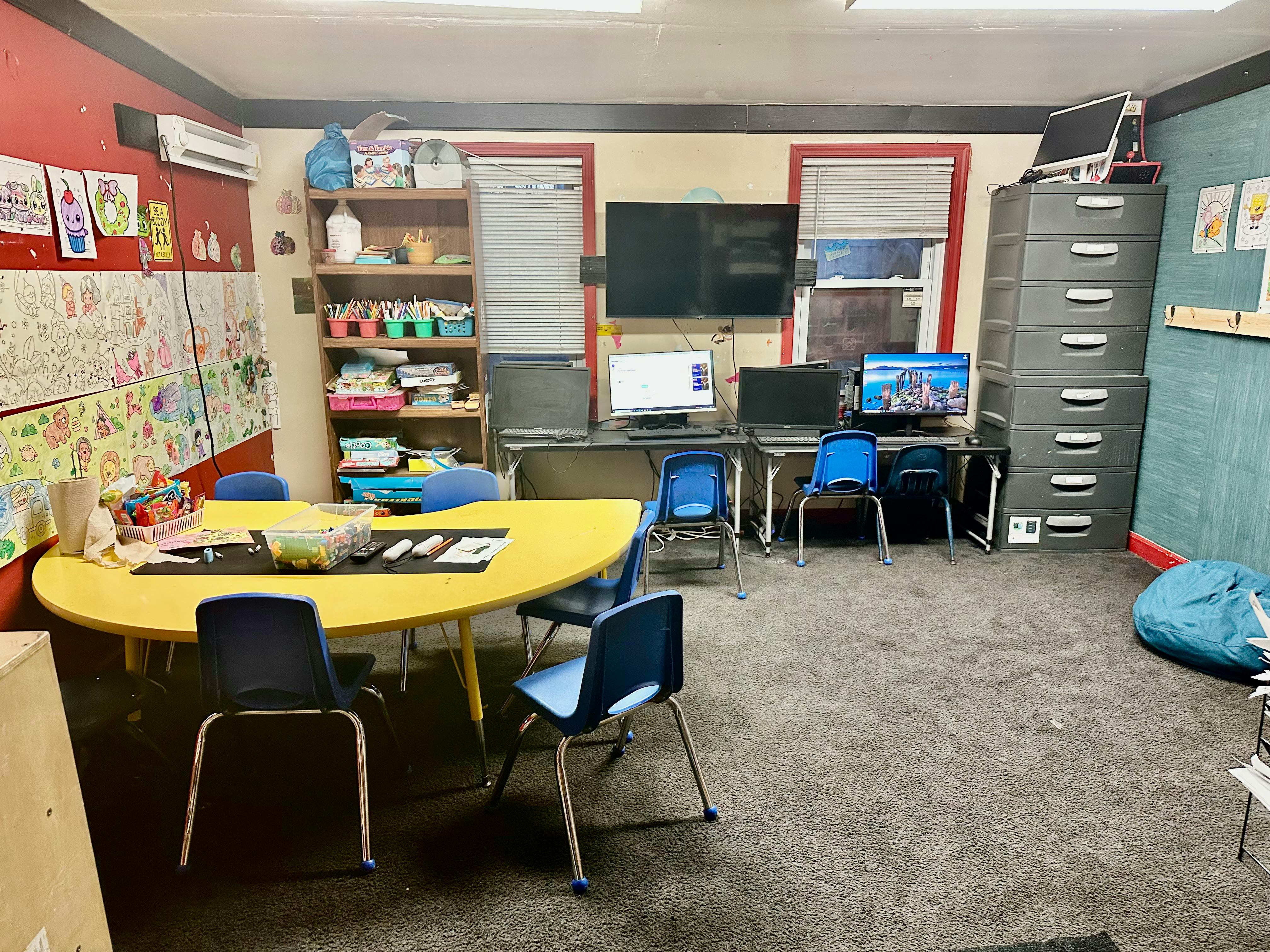 Classroom with yellow tables and blue chairs, computers, and shelves