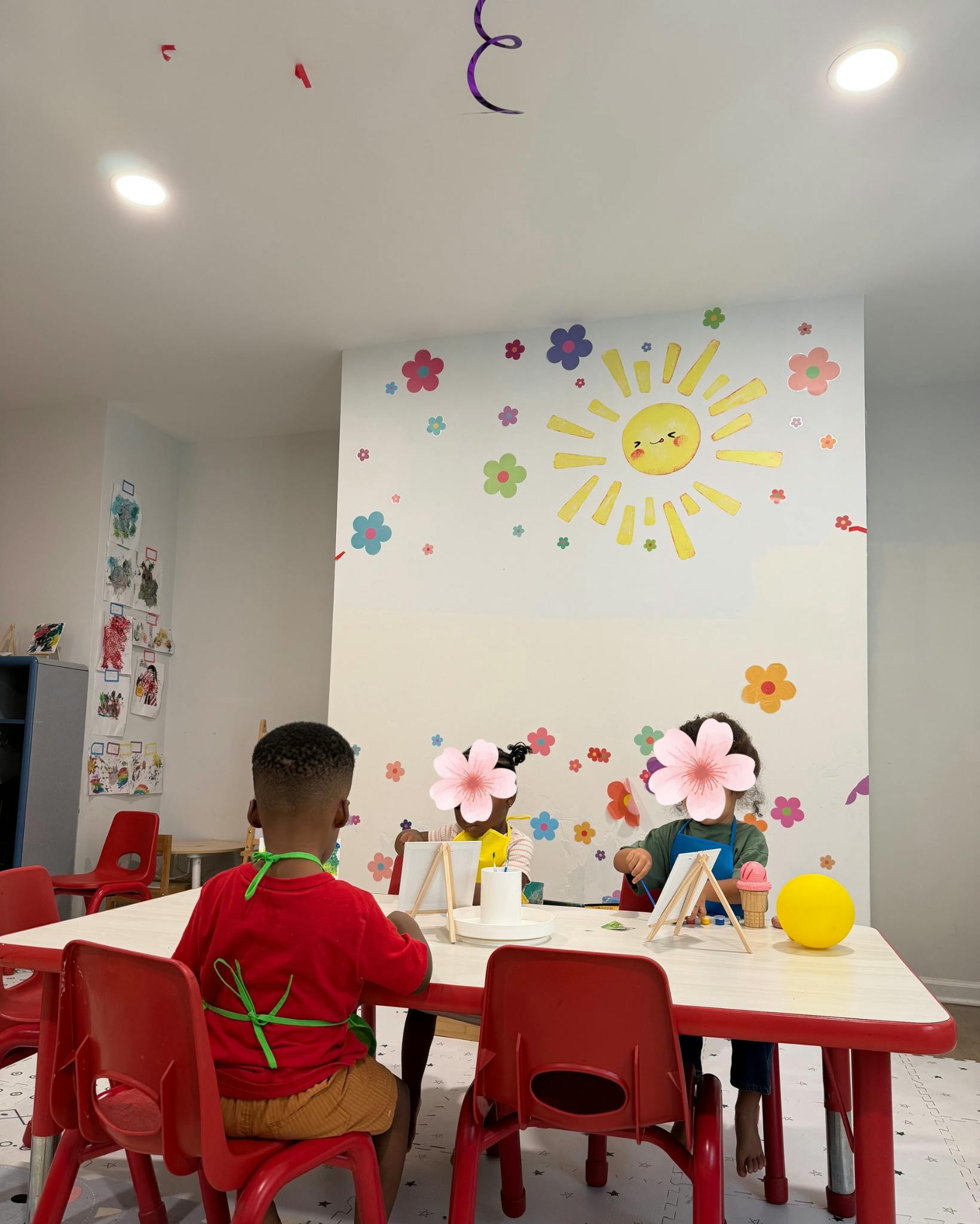 Three children sitting at a table painting on small canvases in a classroom