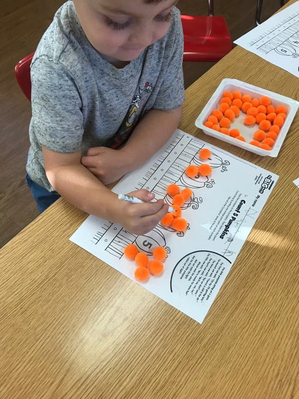 Child counting orange pom-poms on a worksheet in a classroom