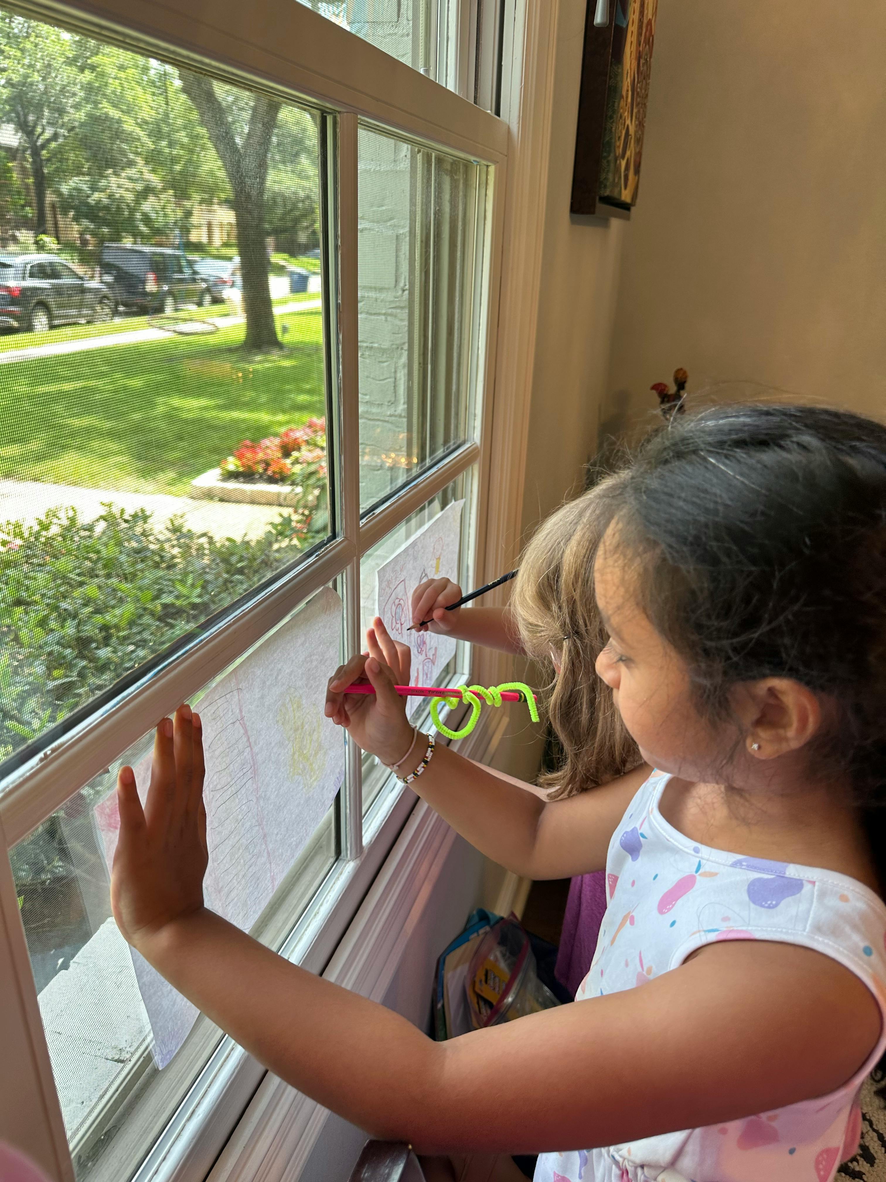 Two children drawing on paper taped to a window