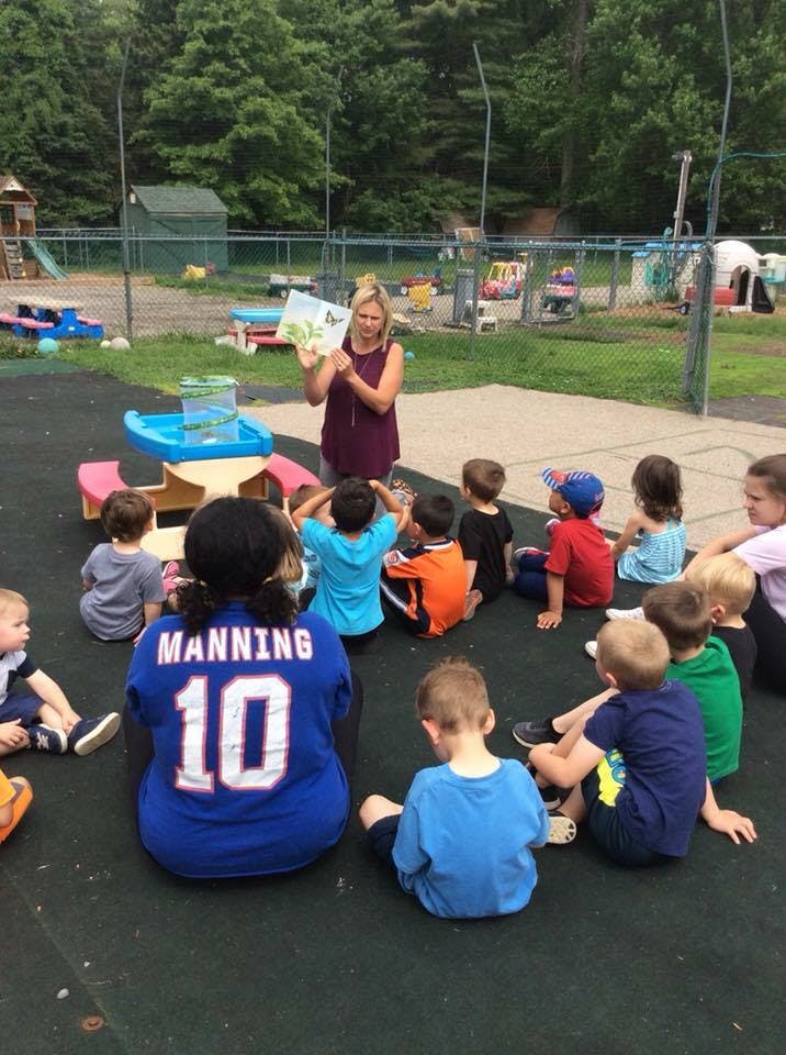 Person reading a book to a group of children sitting on the ground outdoors