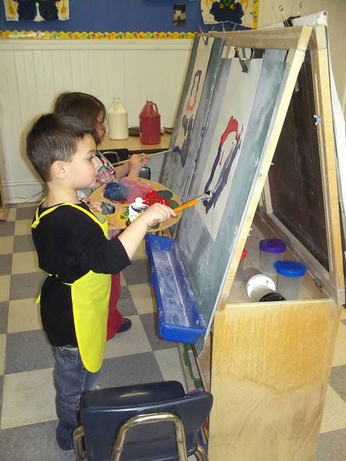 Child in a yellow apron painting at an easel