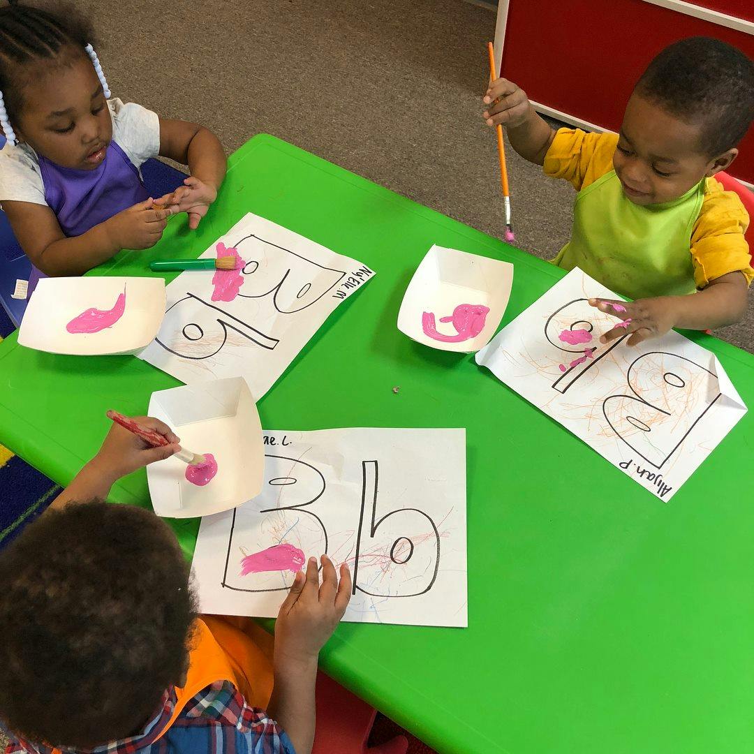Three children painting letters on paper at a green table
