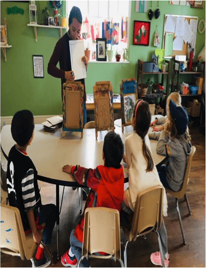Person holding paper and speaking to children seated at a table in a classroom