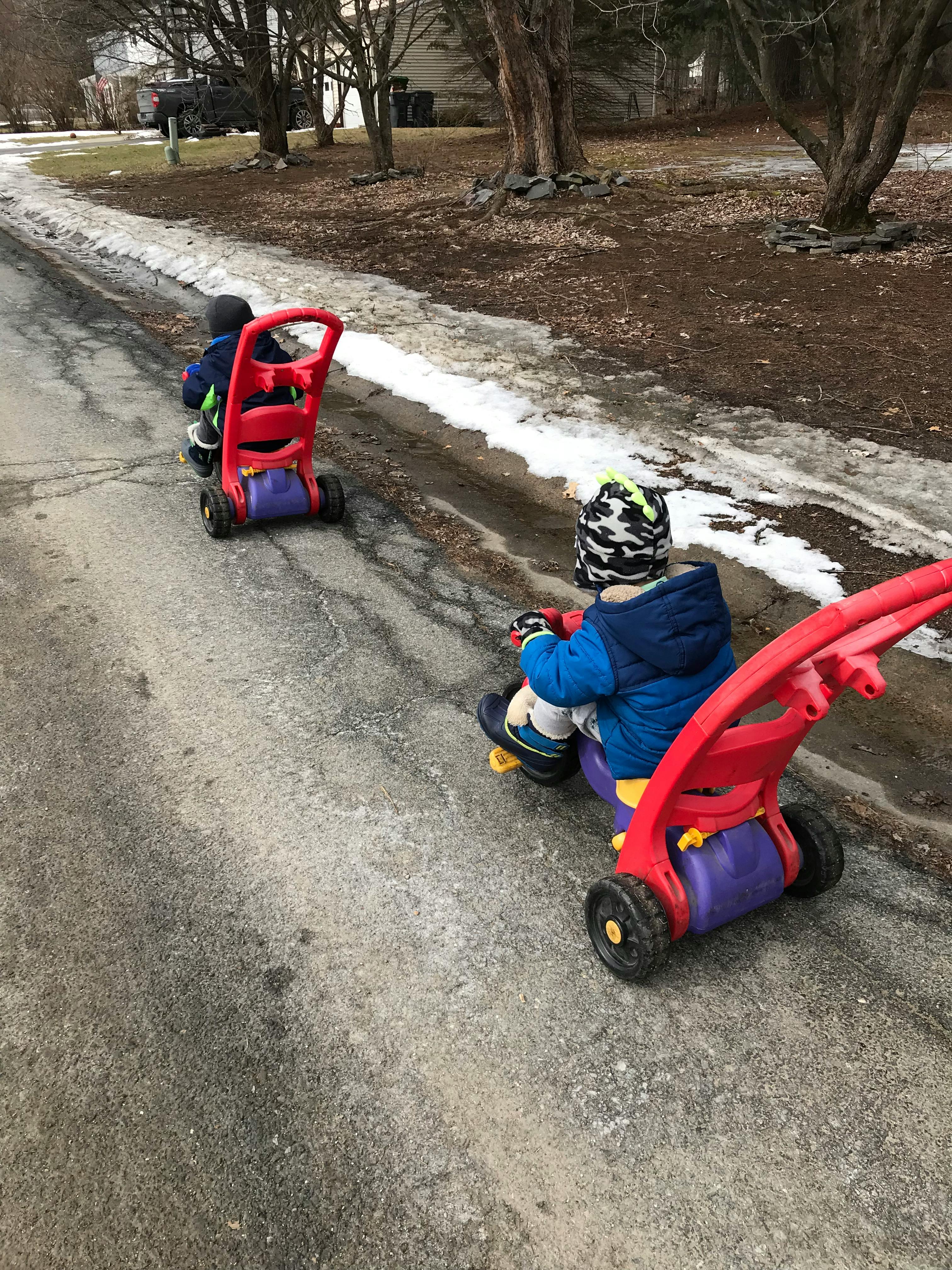 Two children riding toy lawnmowers on a paved surface
