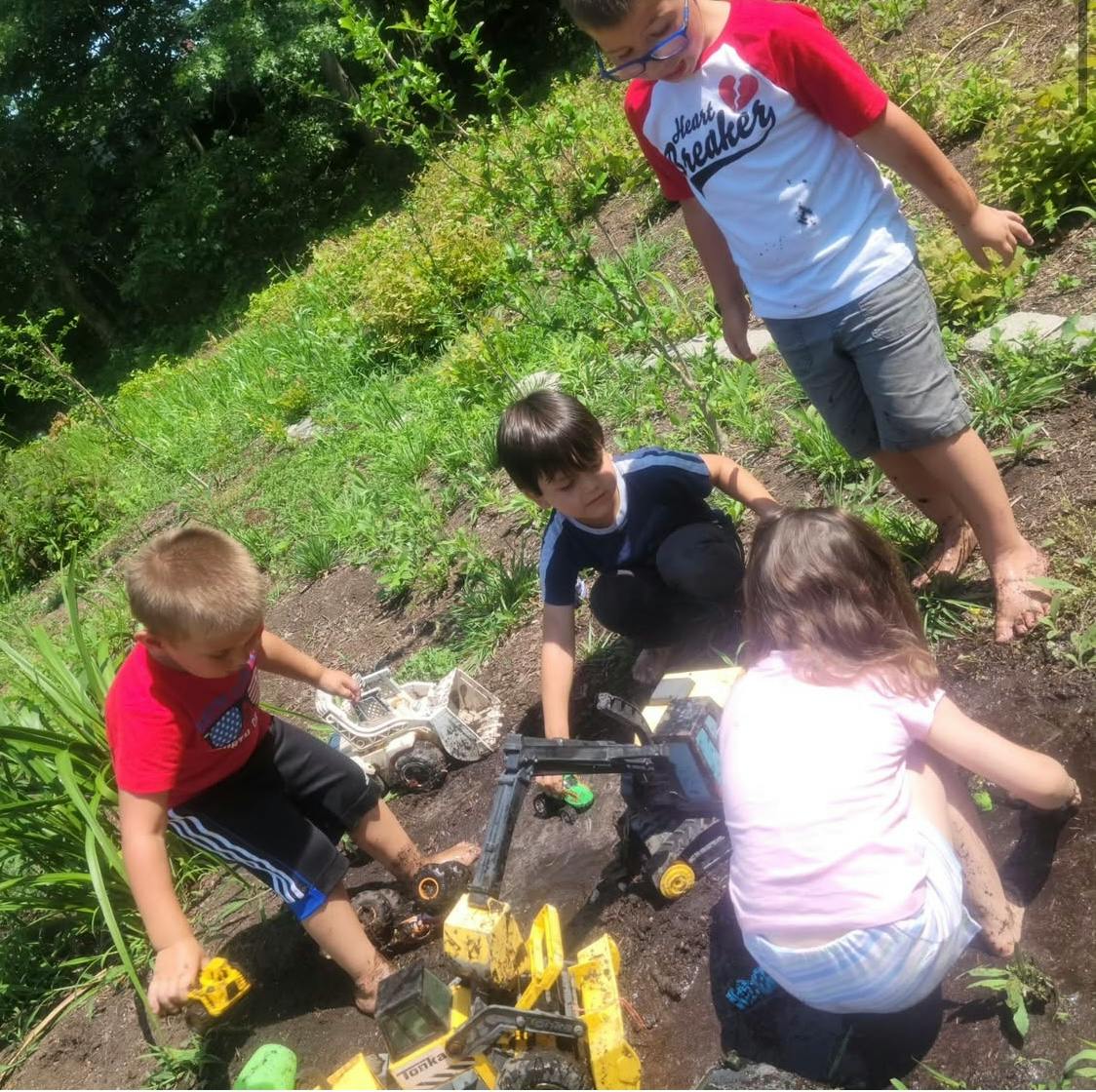 Three children and one teen playing with toy construction vehicles in dirt