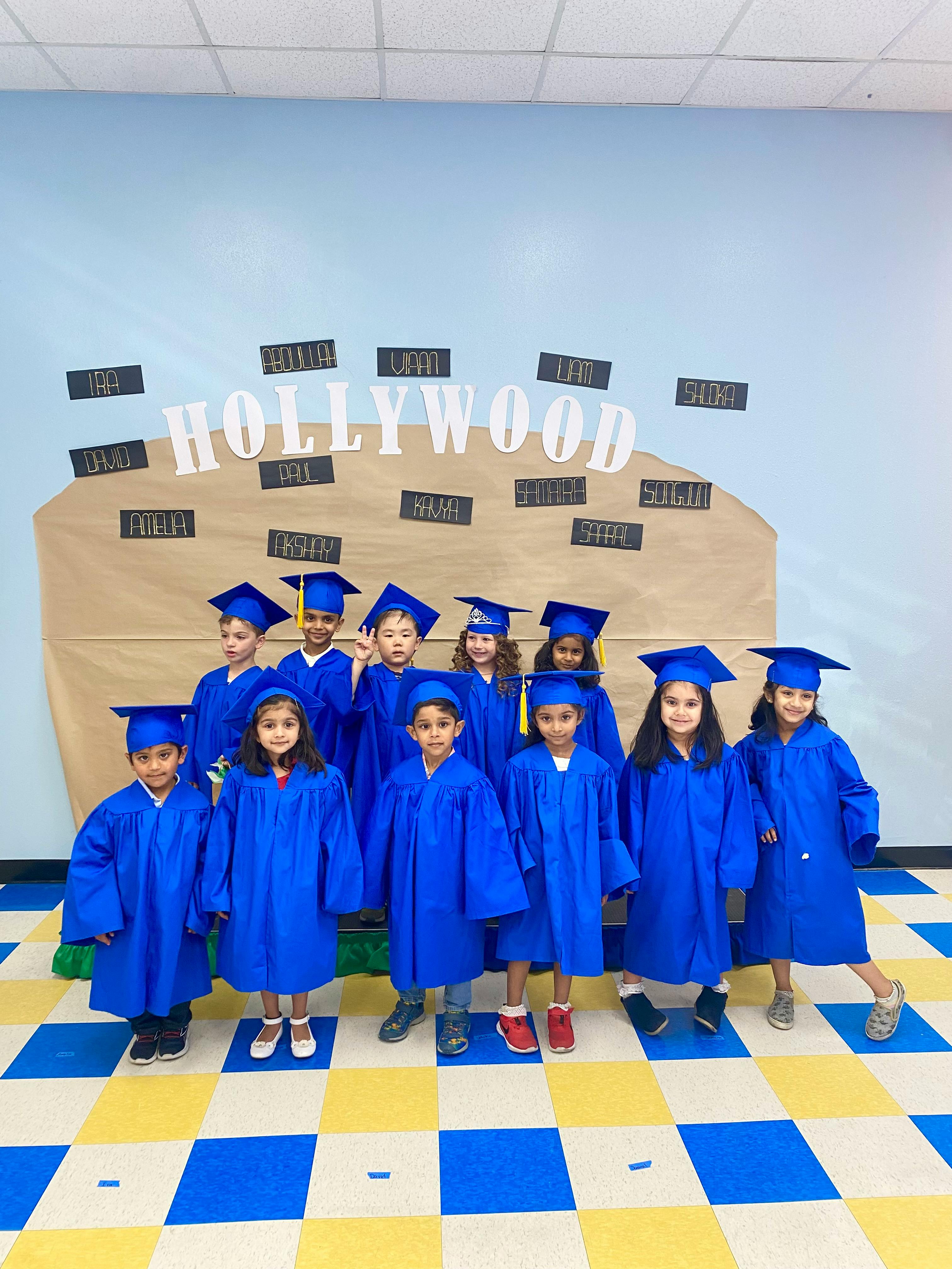 Group of children in blue graduation caps and gowns standing in front of a Hollywood sign