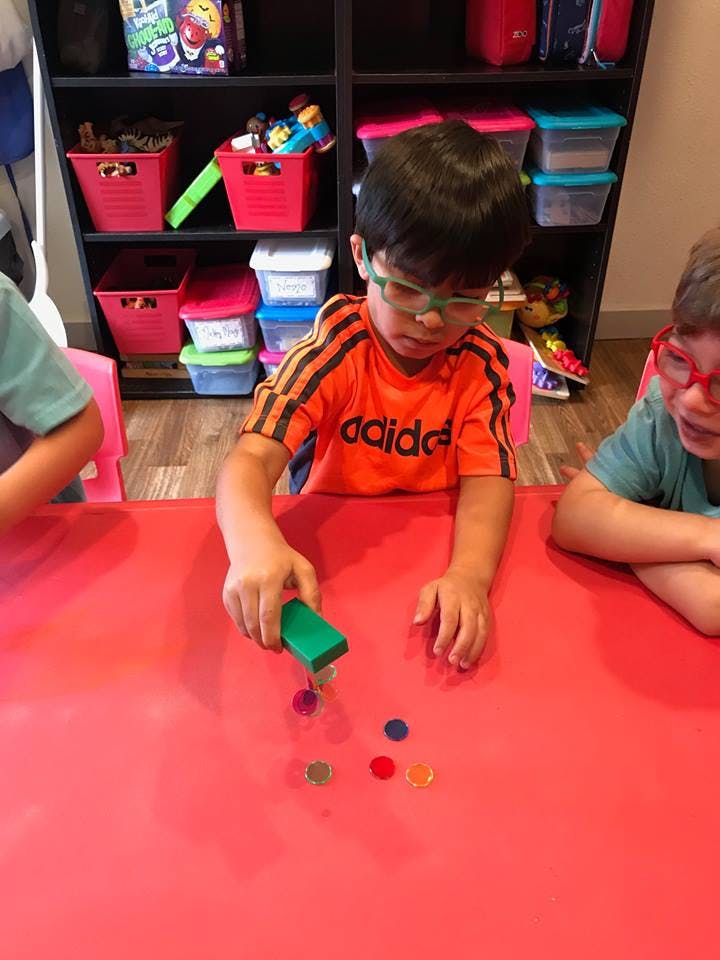 Child in glasses playing with a green block and colorful tokens on a red table