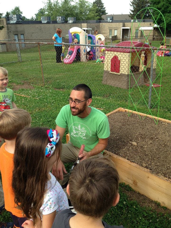 Adult person with glasses talking to children next to a raised garden bed