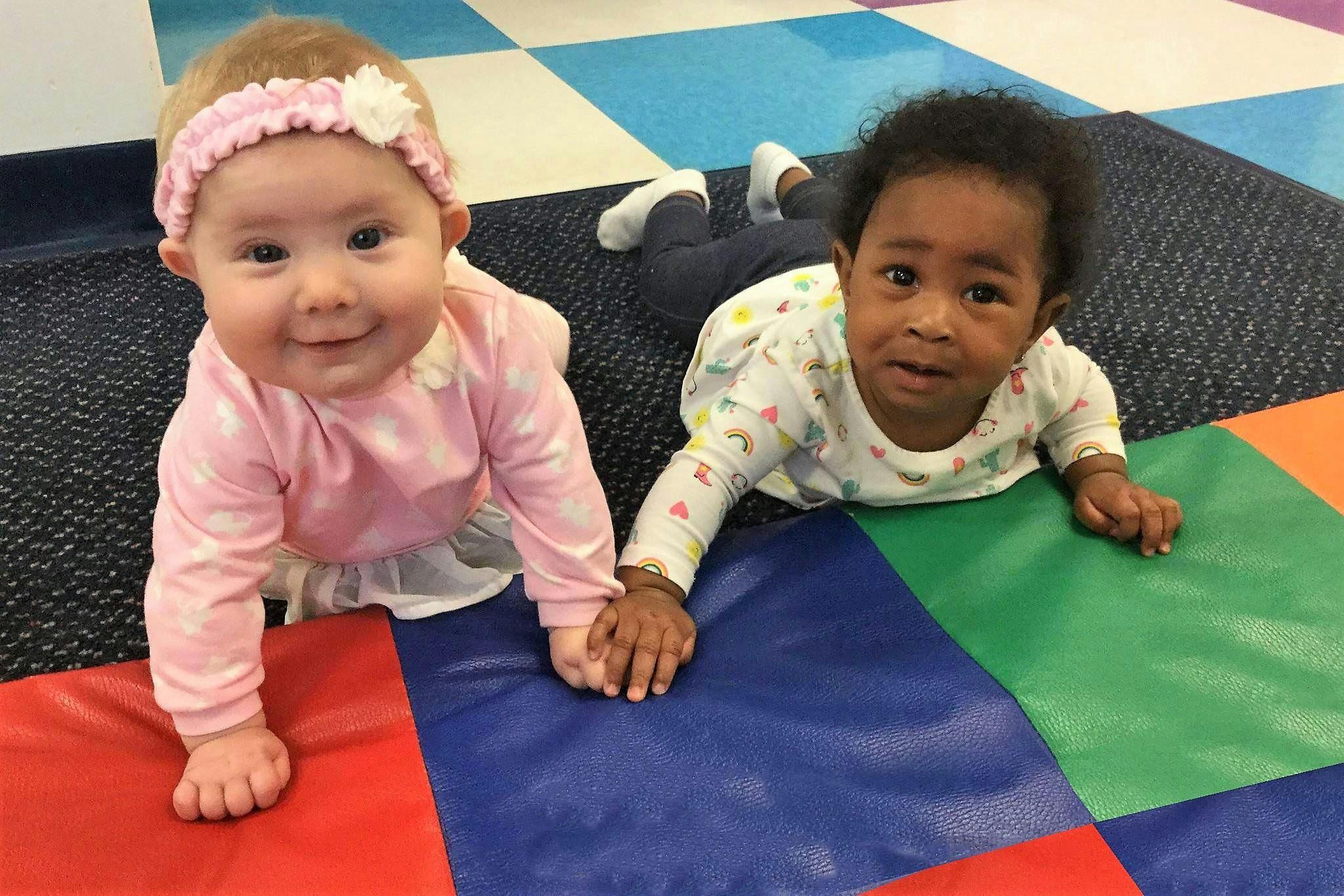 Two babies crawling on a colorful mat, holding hands