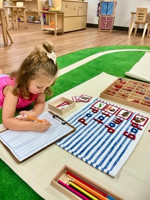 Child writing on a clipboard with letter tiles and picture cards in a classroom