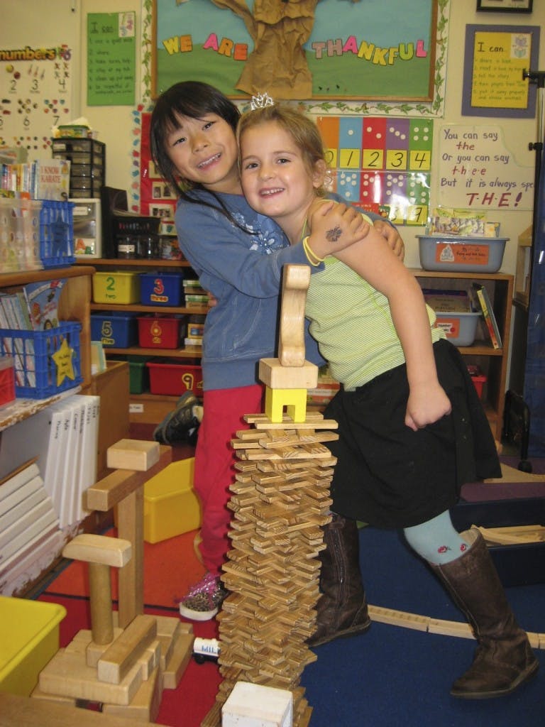 Two children hugging next to a tall block tower in a classroom