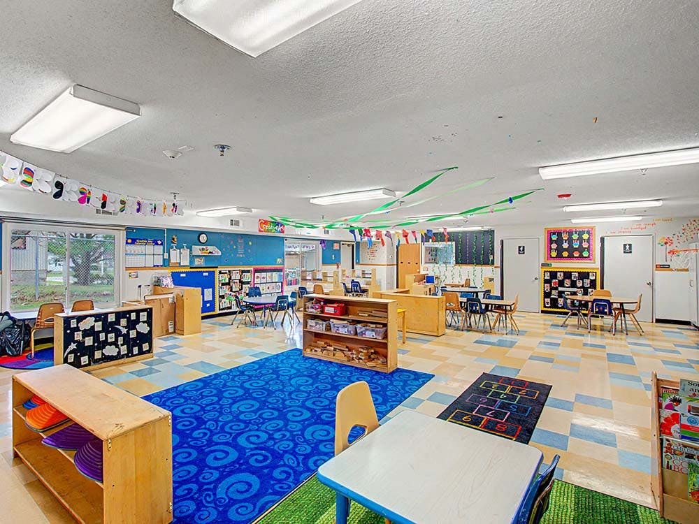 An empty classroom with small tables and chairs, colorful rugs, and decorations