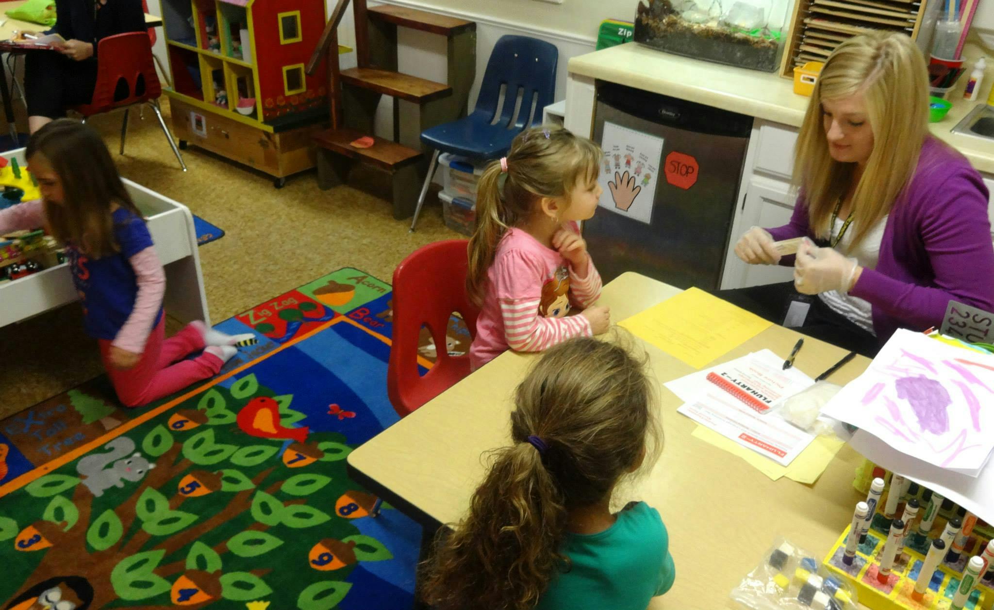 Adult and two children at a table in a classroom, another child on the floor