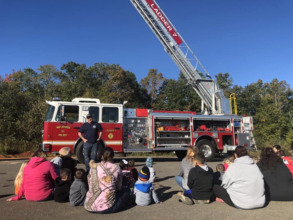 People and children sitting on the ground in front of a fire truck with a ladder extended