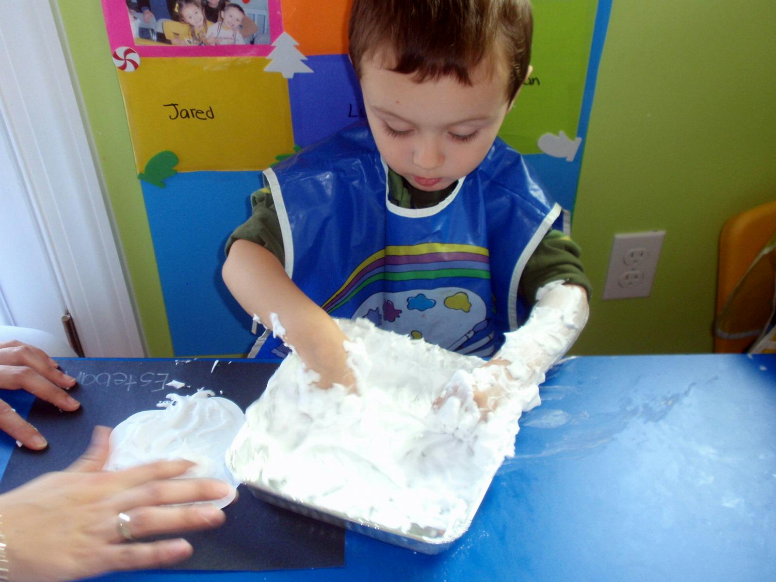 Child in a blue smock playing with white foam in a tray, adult hand nearby