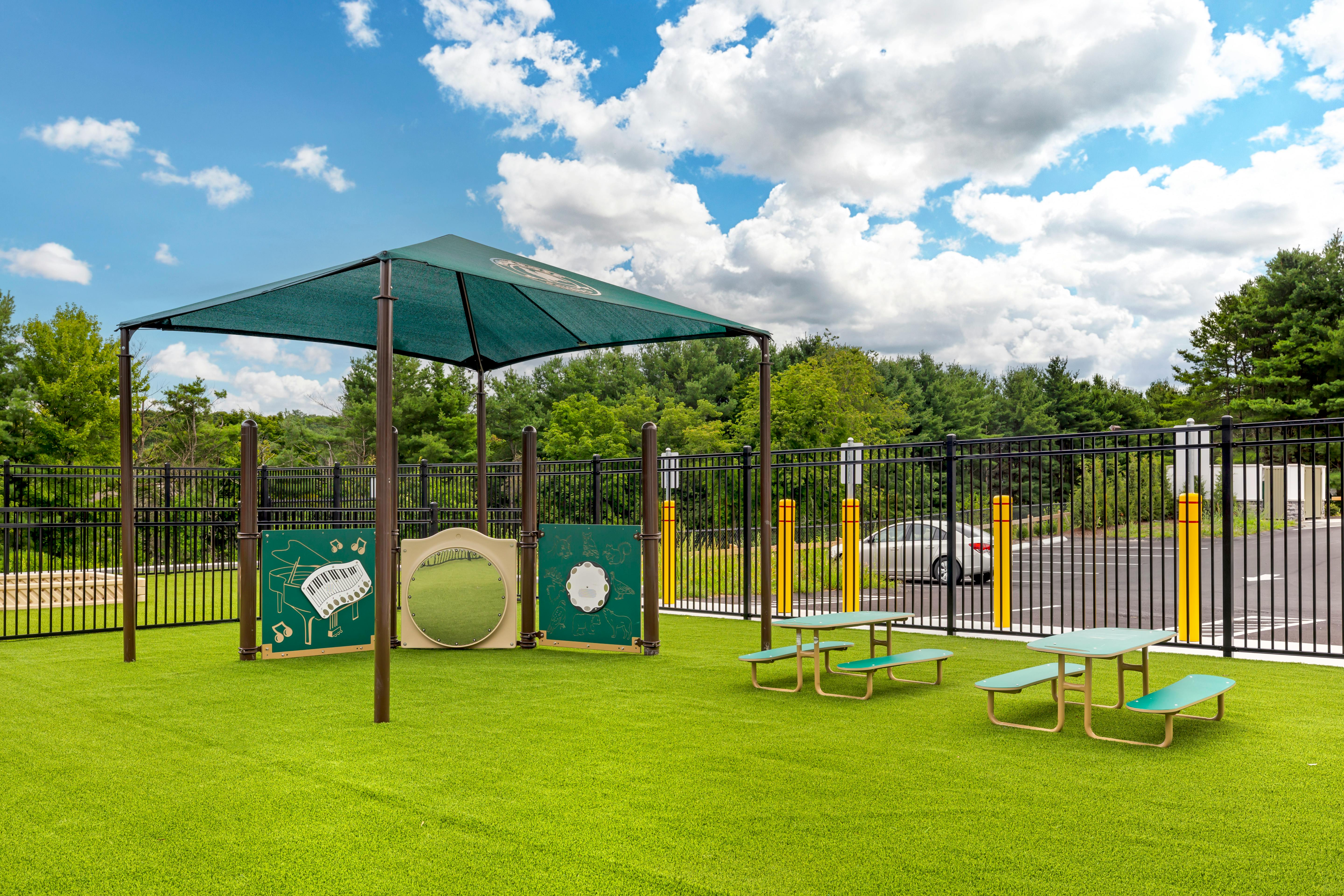 Playground with green turf, a shaded structure, activity panels, and small picnic tables