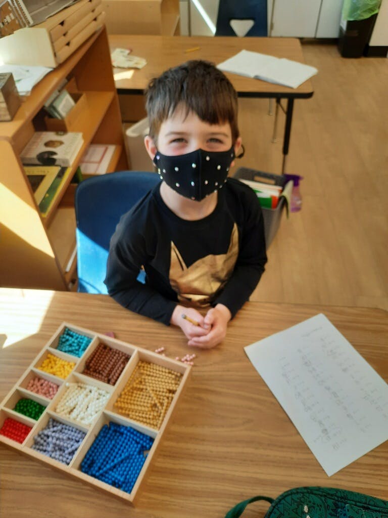 Child in a mask at a desk with a tray of colorful beads