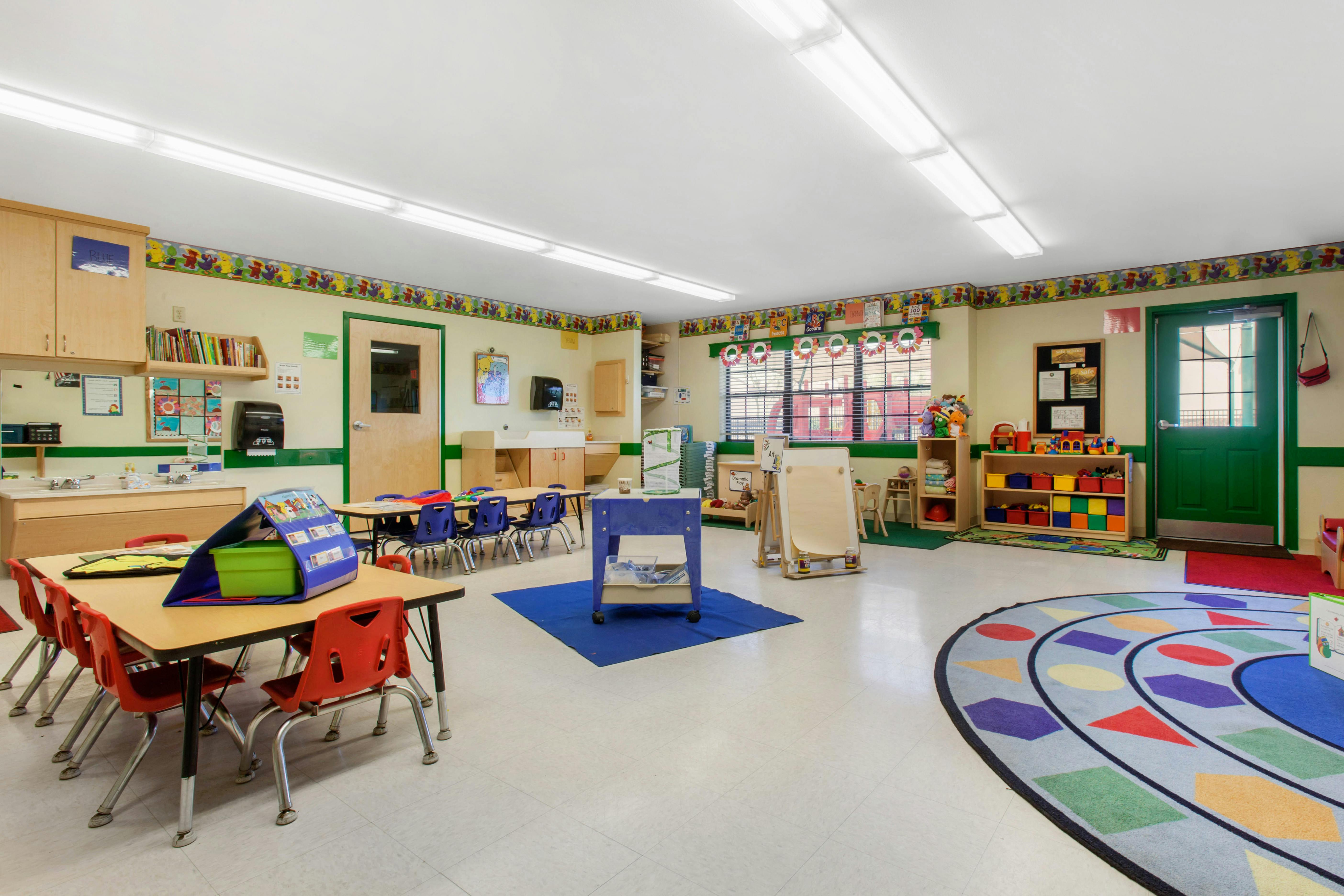Classroom with small tables and chairs, a sink counter, and a colorful rug