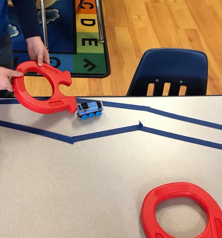 Person holding a red toy, a blue toy train on a table with blue tape