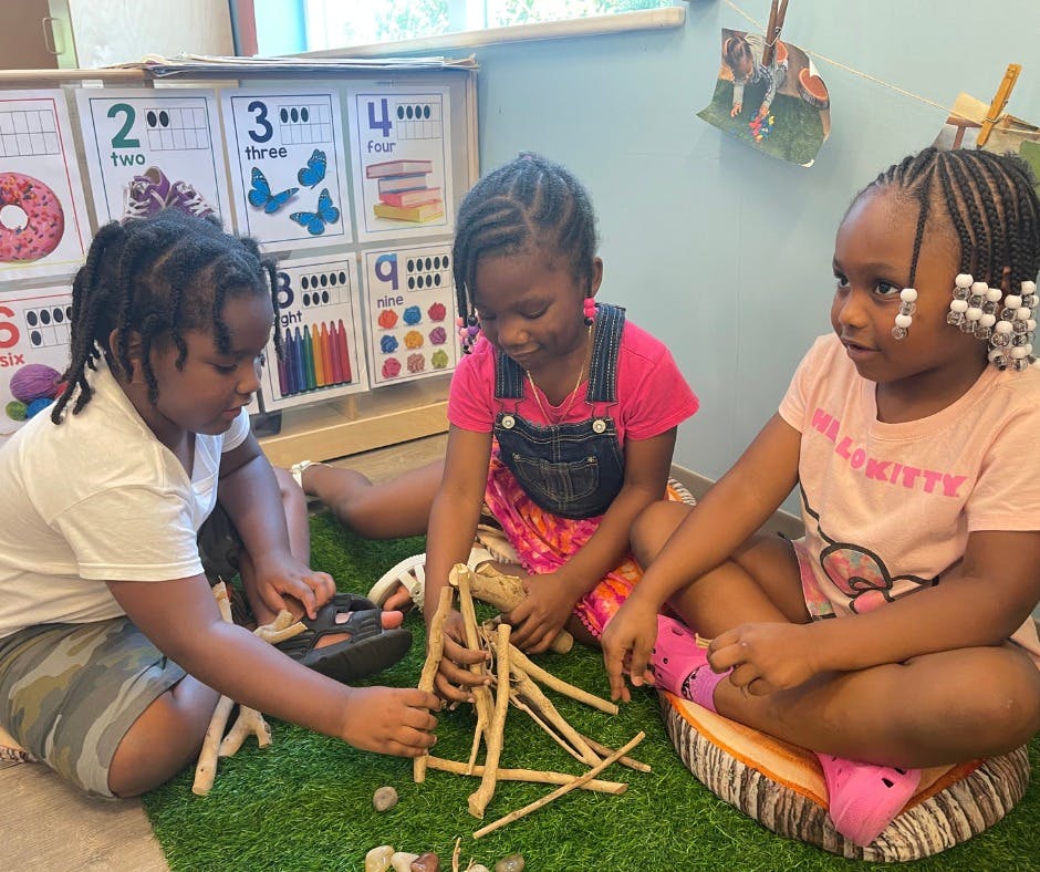 Three children playing with wooden sticks on a green mat