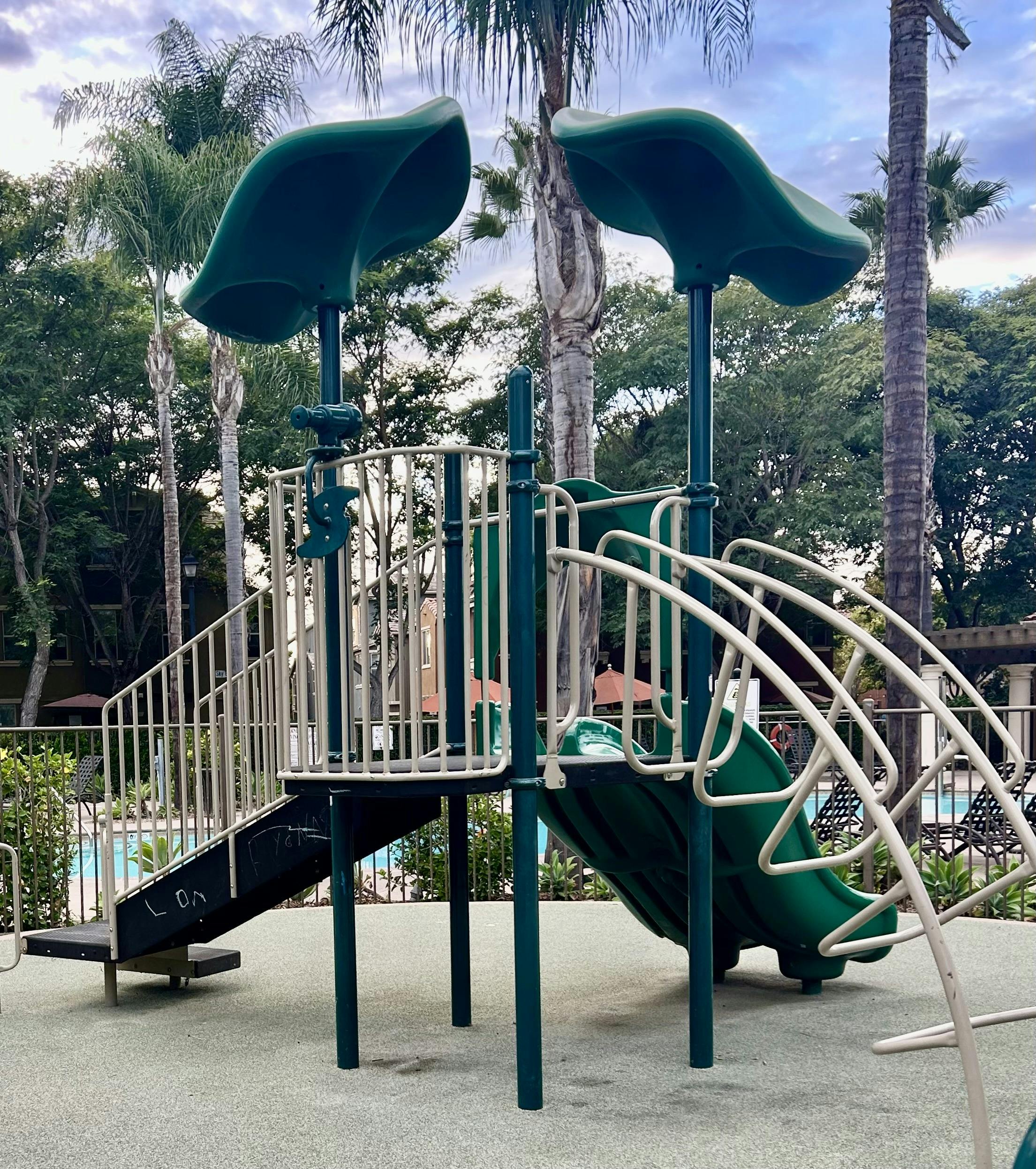 A green and tan playground with slides and ladders next to a swimming pool