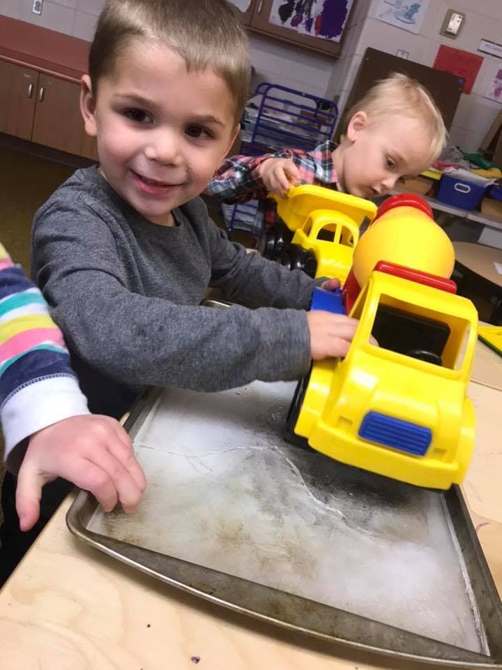 Child playing with a yellow toy truck on a baking sheet with ice