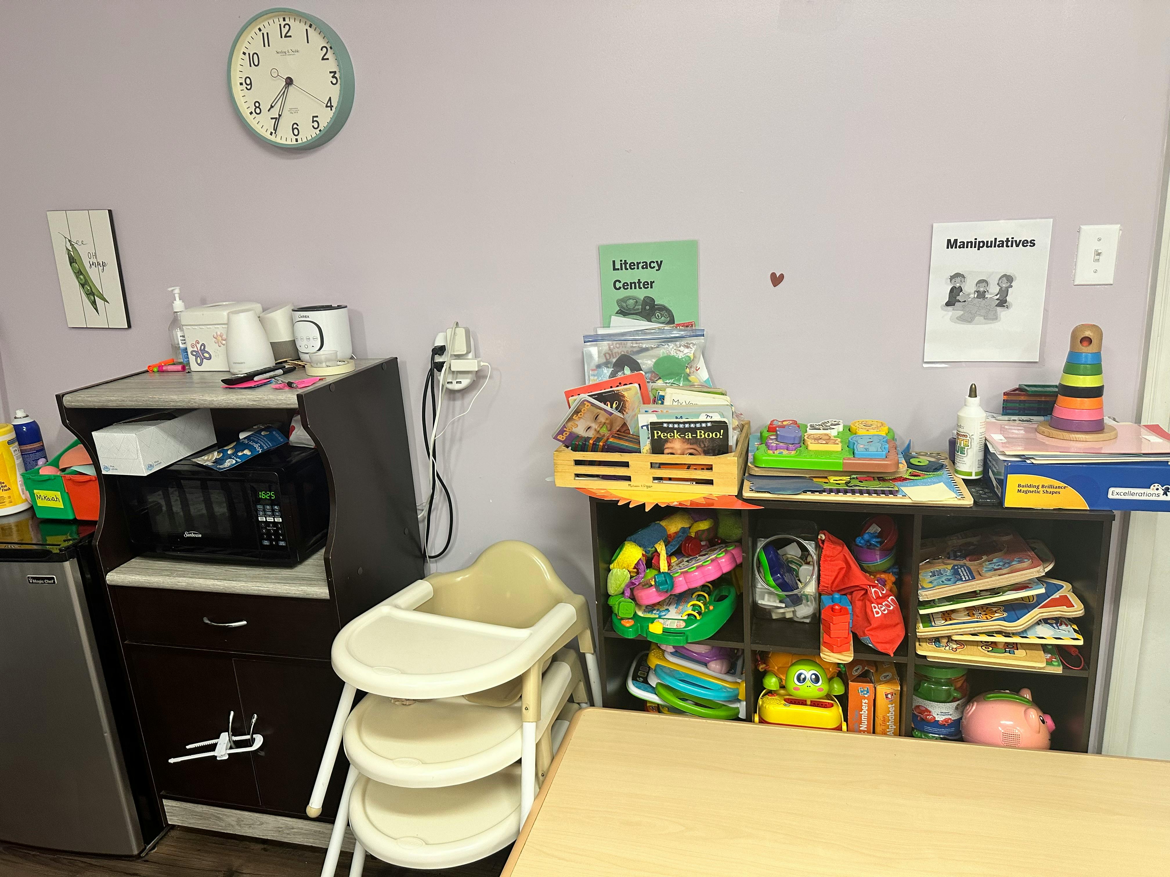 Room with a high chair, shelves of toys, and a small refrigerator
