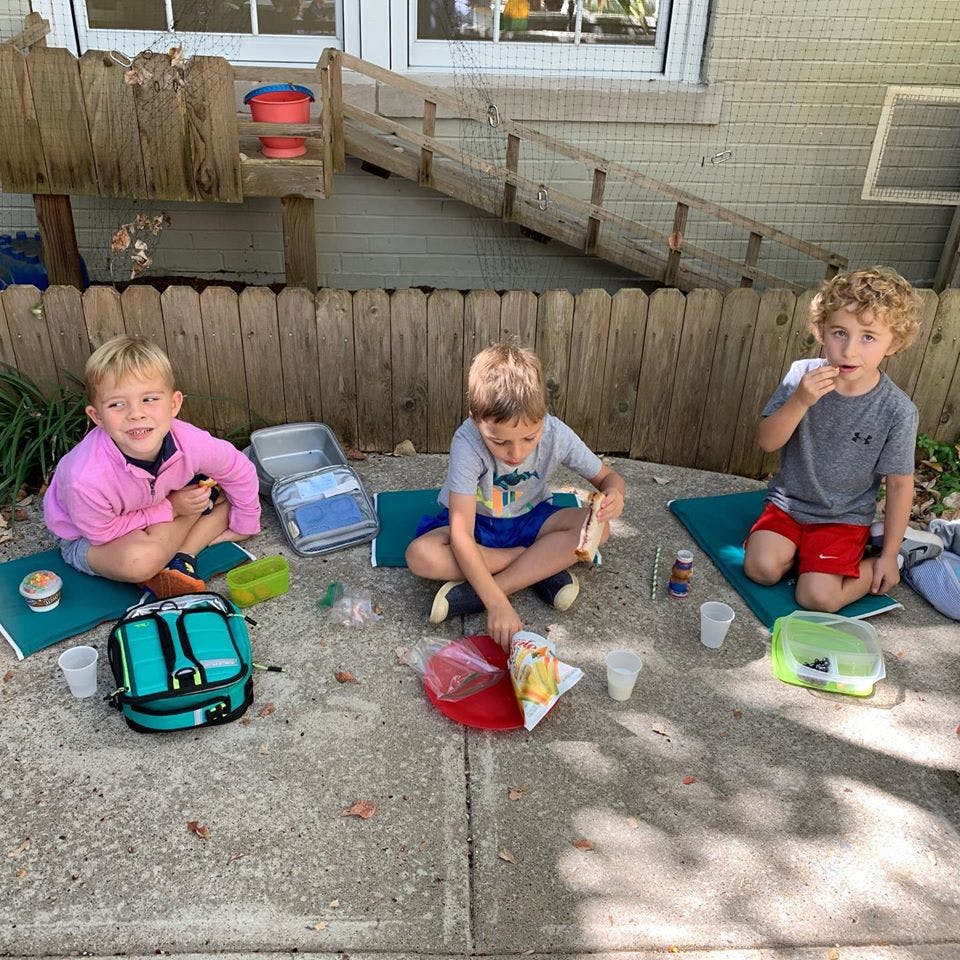 Three children sitting on mats outdoors with lunchboxes and snacks