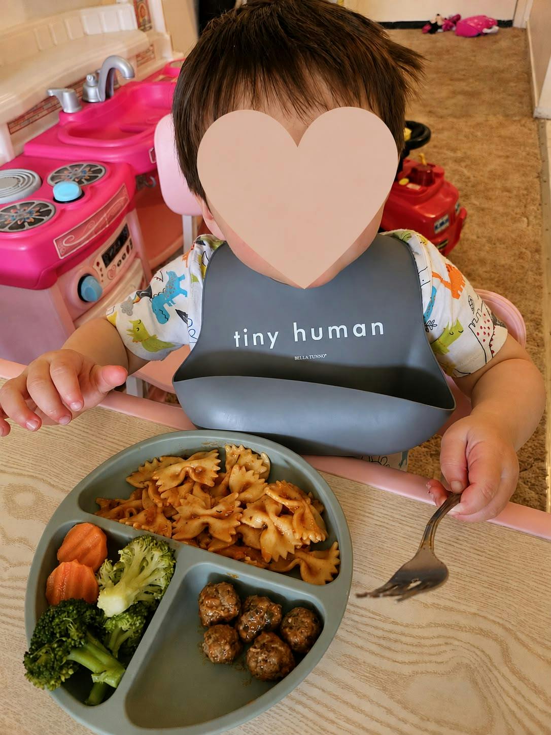 Child wearing a bib eating pasta, meatballs, broccoli, and carrots