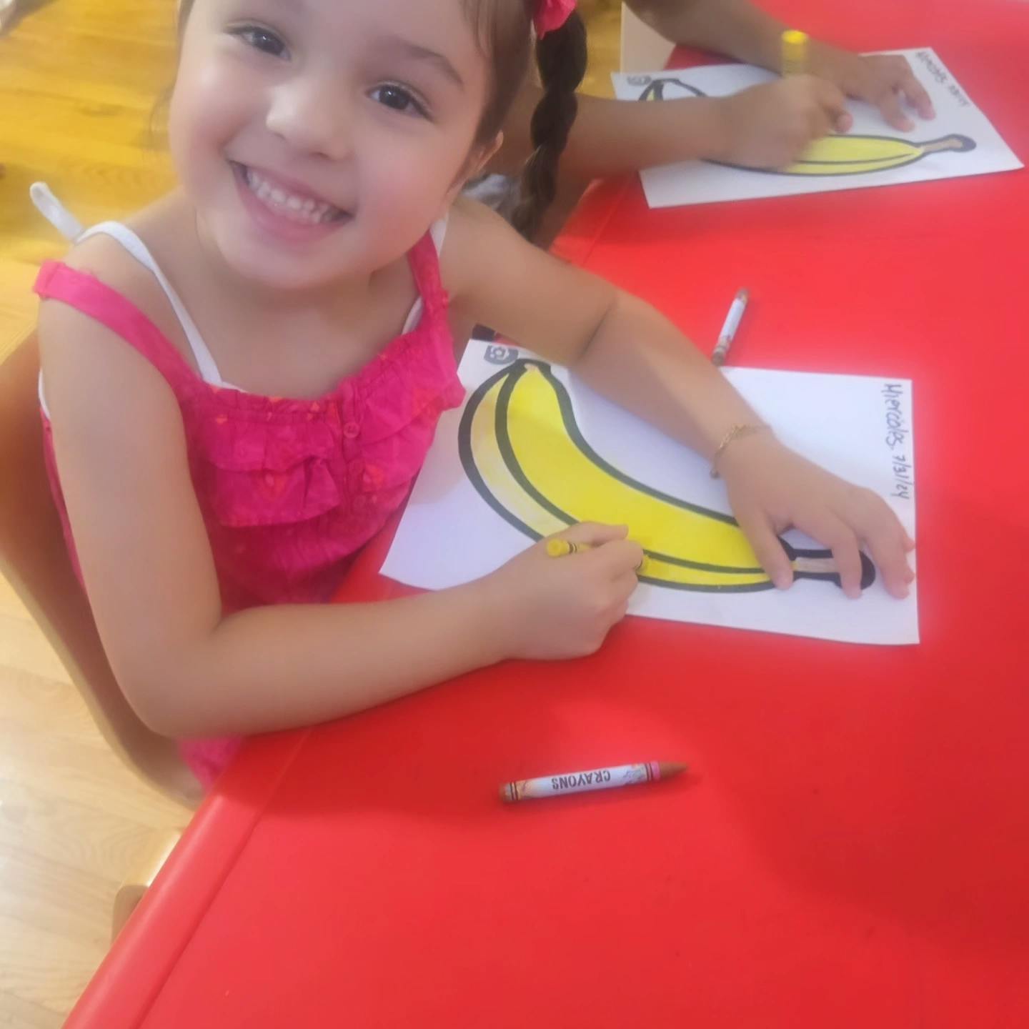 Child coloring a banana drawing at a red table