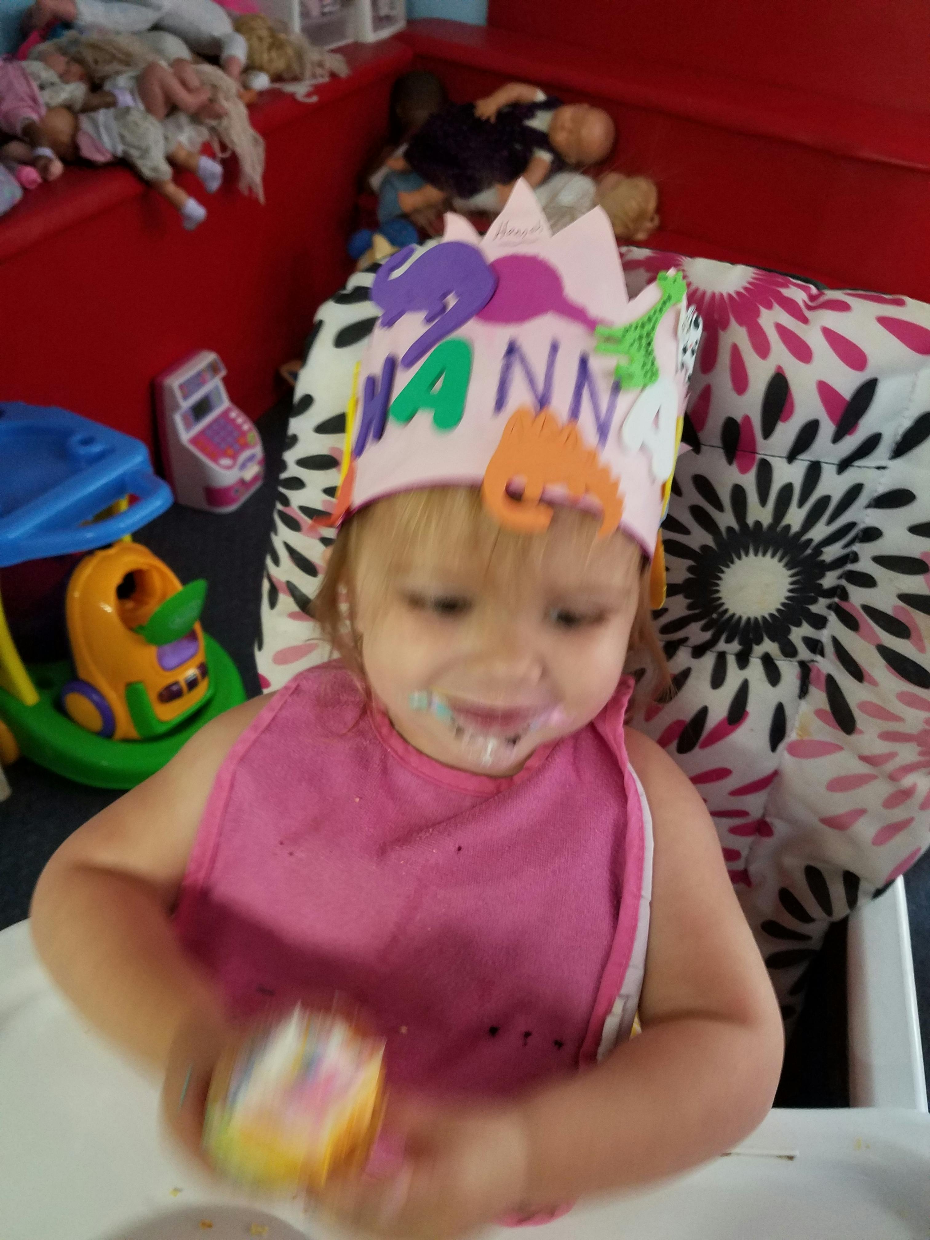 Child wearing a crown and bib, with frosting on their face, holding a cupcake