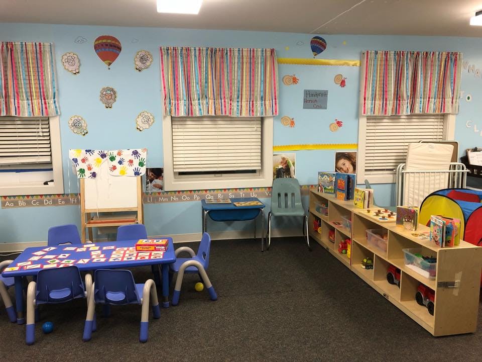 Classroom with small blue tables and chairs, a whiteboard, and shelves with toys