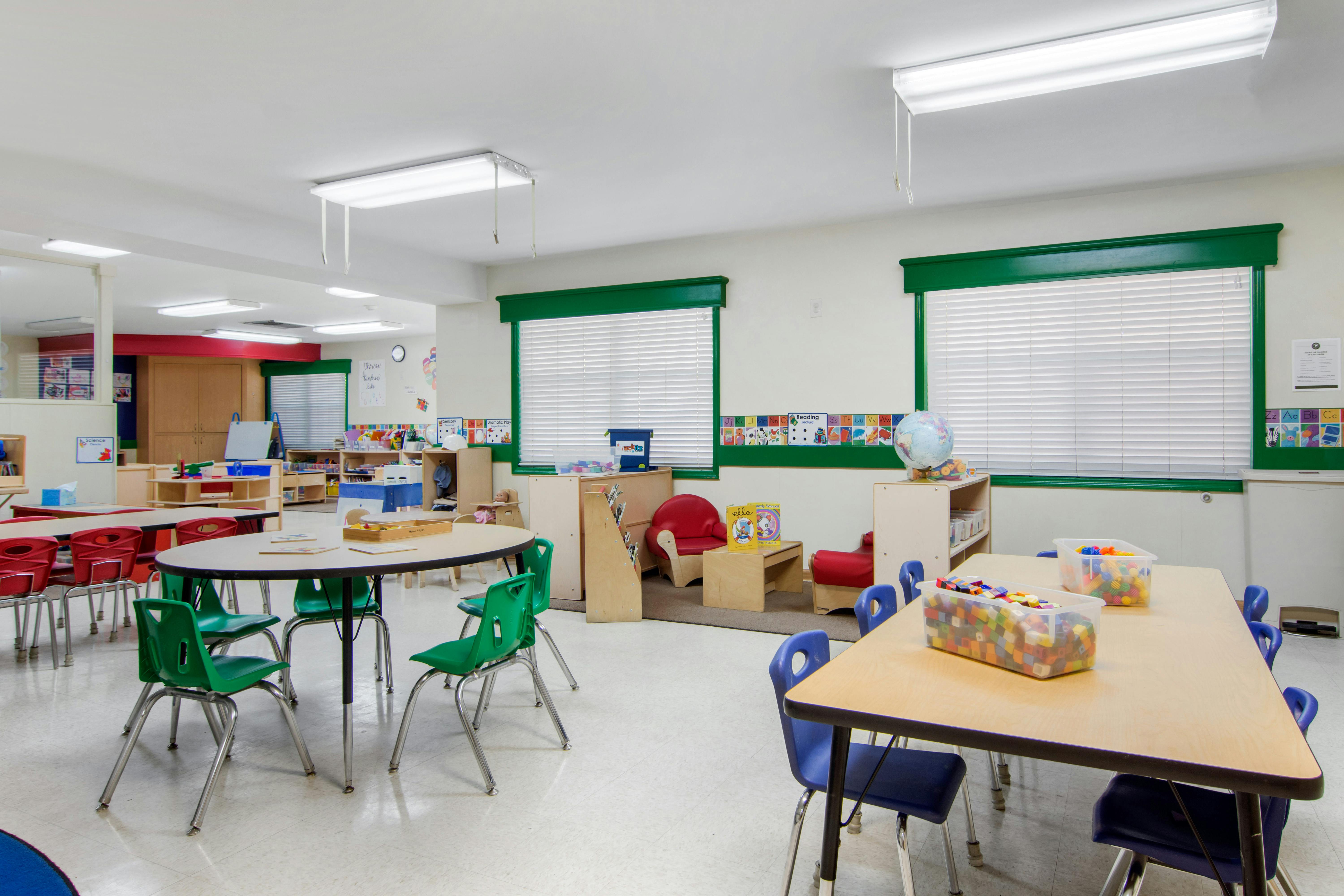 Empty classroom with small tables and chairs, shelves, and windows with blinds