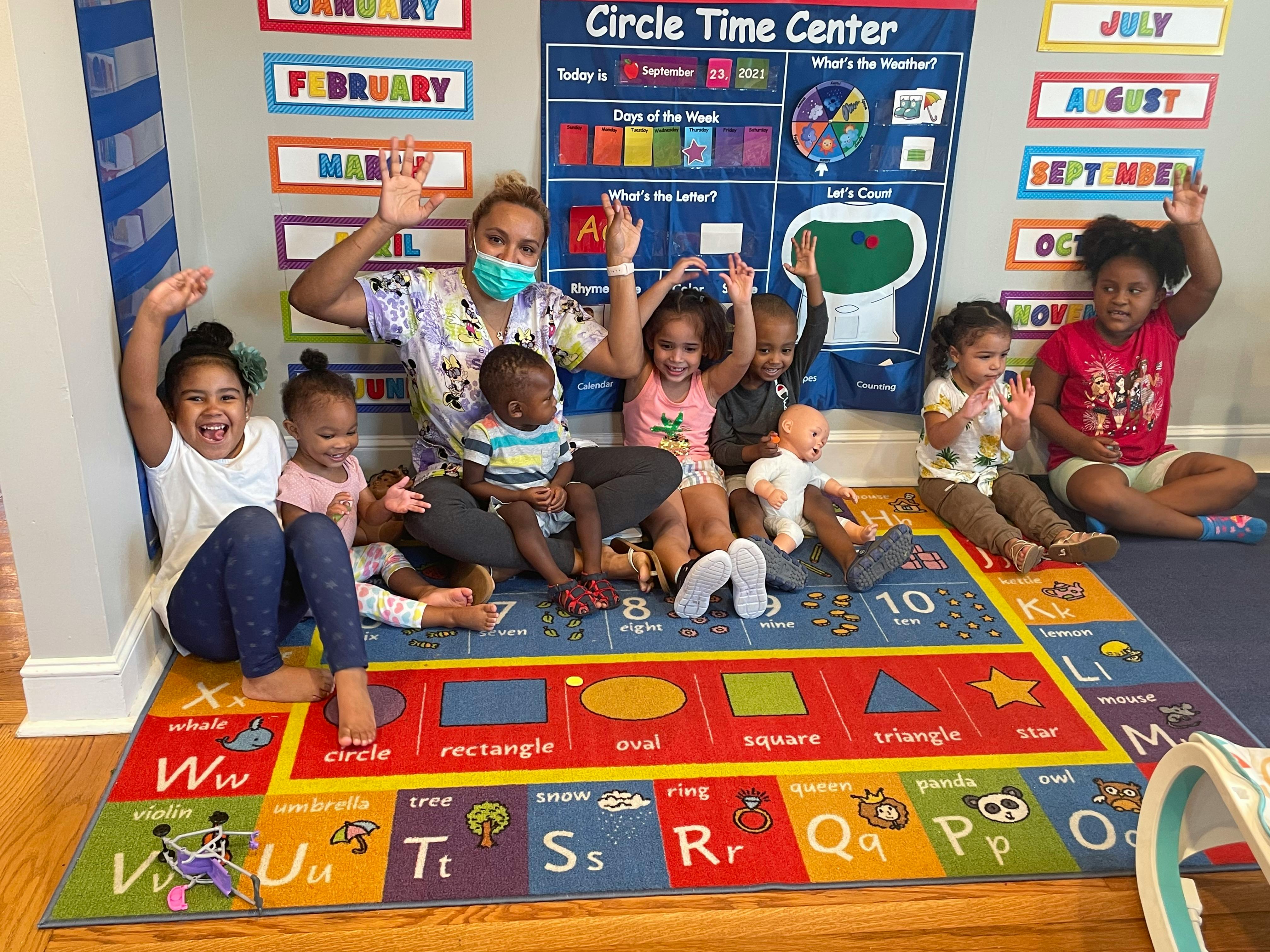 Adult and multiple children sitting on a colorful rug, waving