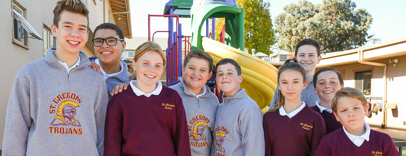 Group of teens and children in school uniform shirts, some with "St. Gregory Trojans" logo