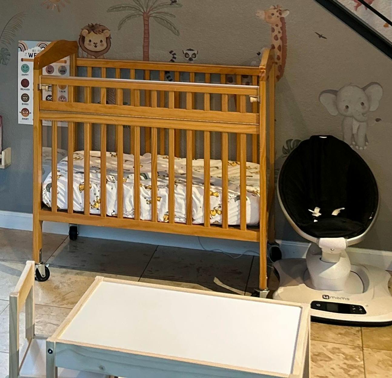 Wooden crib with a patterned mattress next to a baby swing and small table