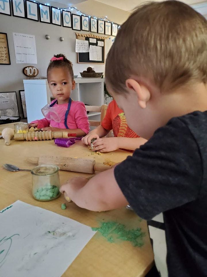 Three children at a table, one looking at the camera, two playing with materials