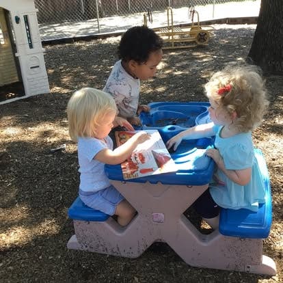 Three children sitting at a small play table looking at a book together